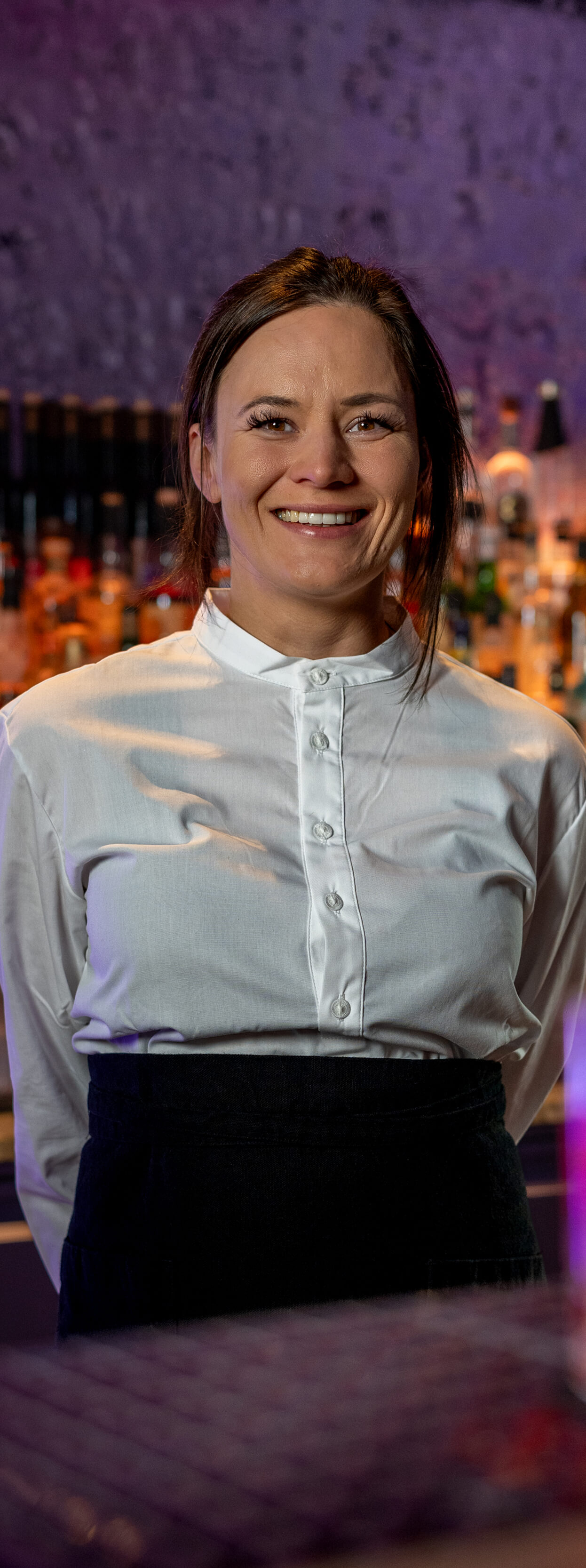 Eine l&auml;chelnde Frau in wei&szlig;em Hemd steht vor einer Bar mit Flaschen im Hintergrund